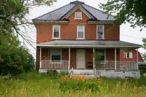 A two-story red brick house with a black roof, featuring a front porch and surrounding greenery. Orange flowers bloom in the foreground, and there is a second structure visible in the background.