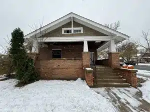 A house with a covered porch, made of brick and wood, surrounded by light snow. There are pumpkin decorations on the steps, and green shrubs on the left side of the image.