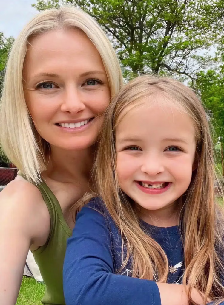 A woman with blonde hair and a sleeveless top sits outdoors, smiling with a young girl who has long hair and wears a blue shirt. They are in a green park-like setting with trees in the background.